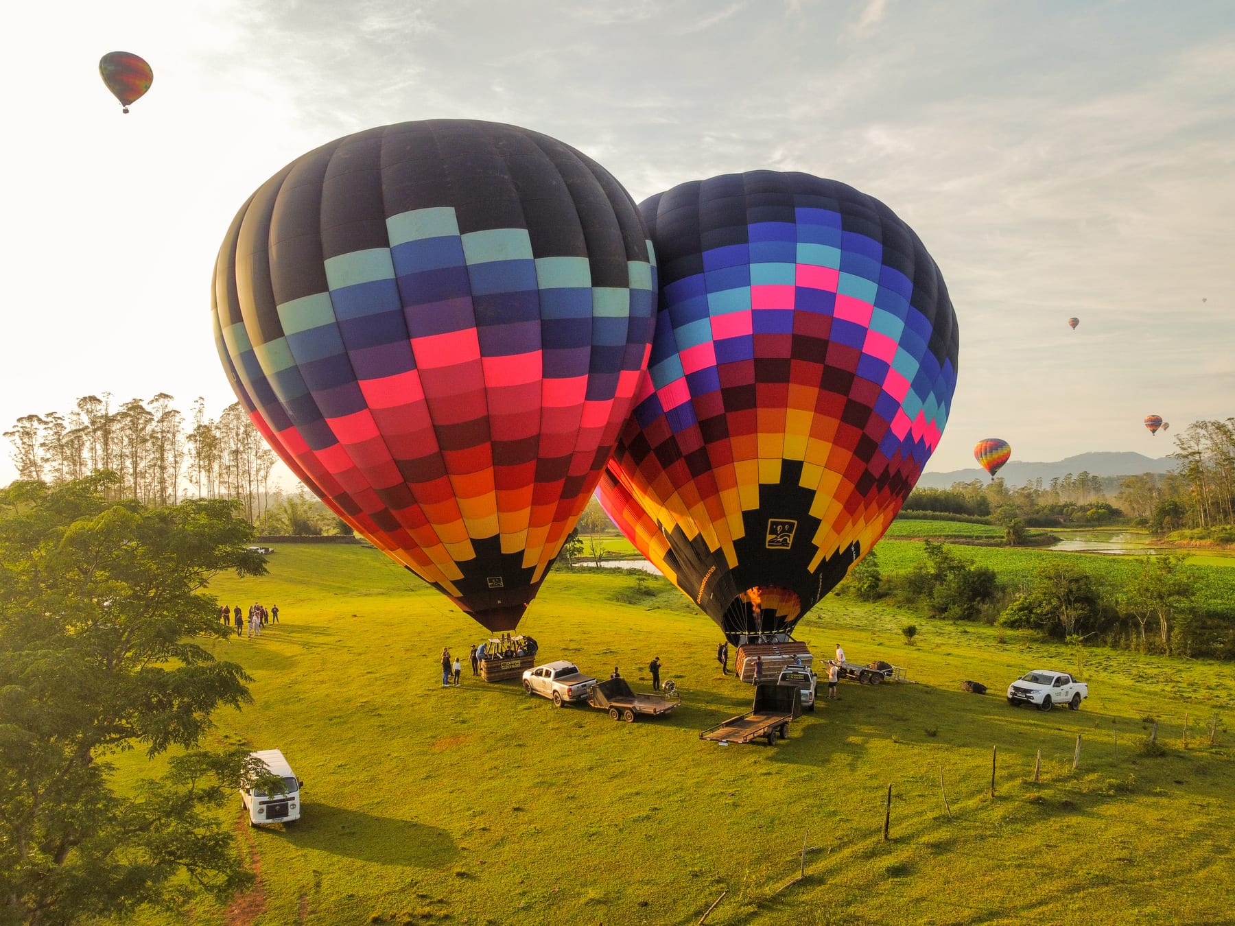 Dois balões coloridos inflados em um campo verde, com a equipe preparando o voo ao amanhecer
